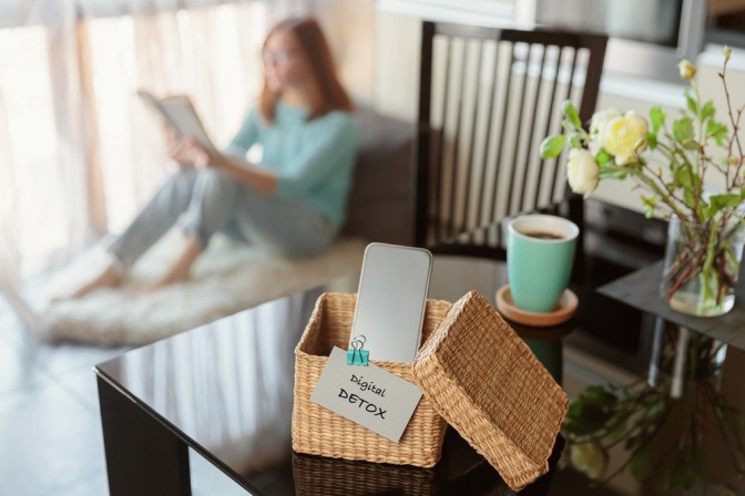 Frau sitzt an Fensterbank und liest ein Buch, ihr Smartphone ist weit weg von ihr.