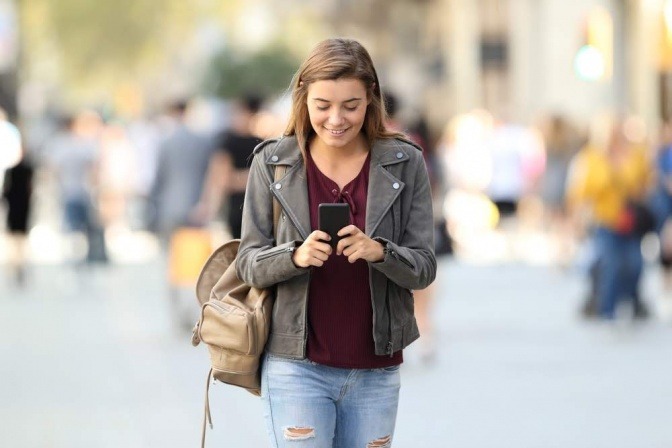 Eine junge Frau läuft in der Gegend herum und schaut dabei lächelnd auf ihr Smartphone.