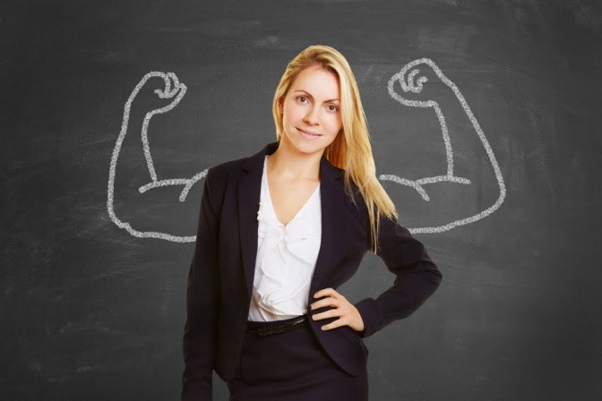 Eine junge Frau im Businessoutfit posiert vor einer Schiefertafel, auf der zwei starke Arme aufgezeichnet sind.