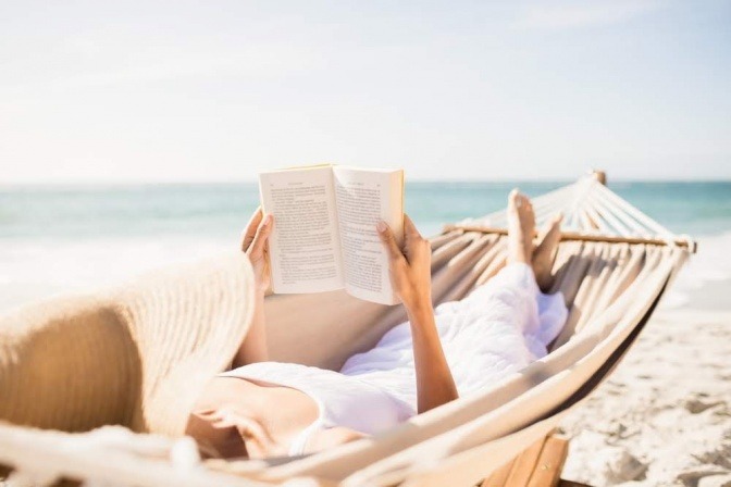 Eine Frau liegt in einer Hängematte am Strand und liest ein Buch. 