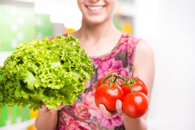 Eine Frau hält einen grünen Salat in der einen und eine Tomatenrispe in der anderen Hand, während sie ihre Arme in Richtung Kamera ausstreckt.