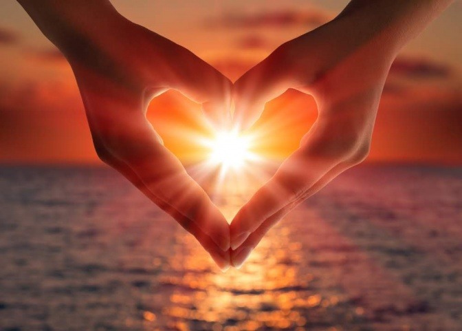 Vor einem romantischen Hintergrund mit einer untergehenden Sonne am Horizont, zeigt das Bild ausschnitthaft die Hände eines Mannes und einer Frau, die gemeinsam ein Herzzeichen bilden.