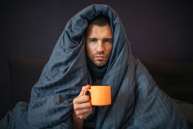 Mann mit Teetasse sitzt auf dem Sofa mit einer Decke über dem Kopf