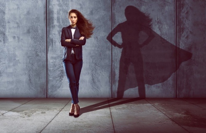 Eine Frau steht vor einer Wand, ihr Schatten zeigt die Silhouette einer Superheldin mit Umhang.