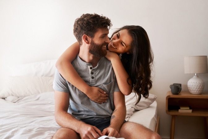 Frau umarmt auf Bett sitzenden Mann und lächelt