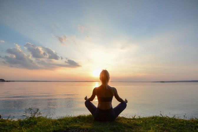 Eine Frau im Schneidersitz meditiert beim Sonnenaufgang draußen am Meer.