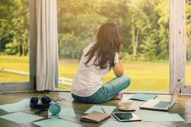 Eine Frau sitzt im Schneidersitz in einer Art Wintergarten und blickt durch die Fenster ins Grüne. Um sie herum liegen diverse Arbeitsmaterialien wie Schreibblock, Tablet, Stifte und mehr.