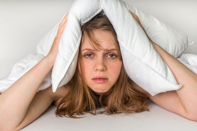 Eine Frau hat offenbar Schlafprobleme und hält sich ein Kissen über den Kopf, während sie mit einem erschöpften Blick aus dem Bett schaut.