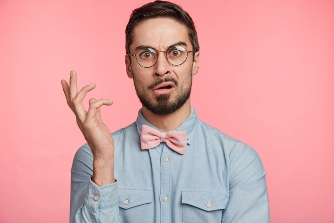 Körpersprache, Männer. Berührungen - Porträt eines unzufriedenen Mannes, der eine runde Brille trägt, einen mürrischen Blick hat, mit der Hand gestikuliert und sich über etwas wundert, posiert vor einem rosa Studiohintergrund.