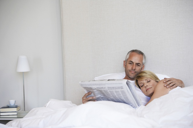 Paar liegt im Bett, Mann liest Zeitung