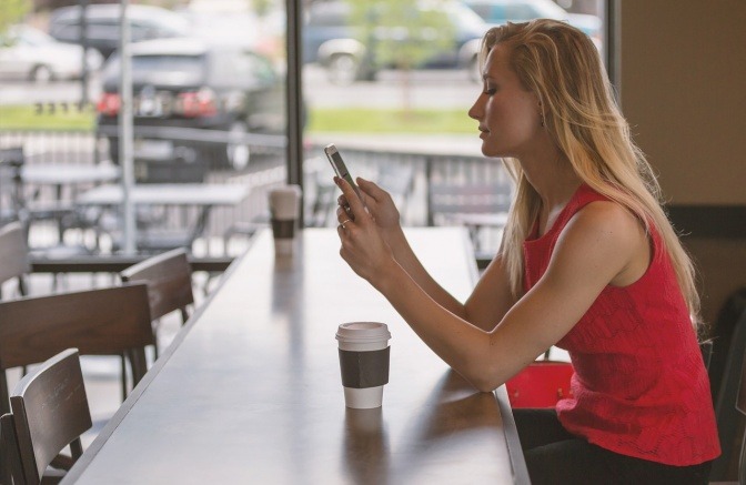 Eine junge blonde Frau sitzt an einer Theke und blickt auf ihr Smartphone.