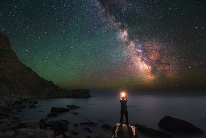 Ein Mann steht im Dunkeln auf einem Stein in einer Bucht und hält eine stark leuchtende Kugel in den Händen, die er in den Himmel streckt.