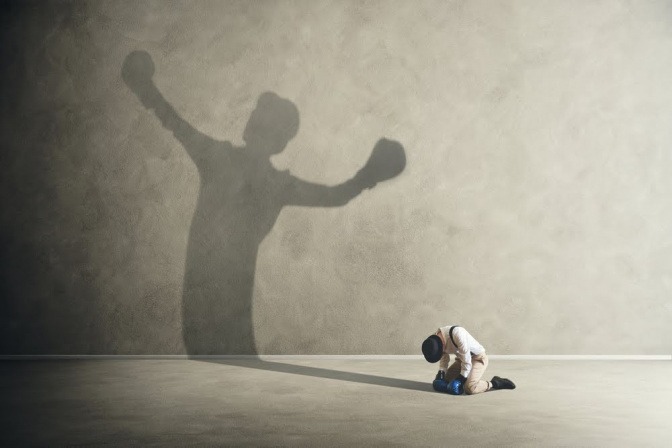 Ein Boxer kniet niedergeschlagen auf dem Boden. Sein Schatten an der Wand reckt jedoch triumphierend die Hände in die Luft.