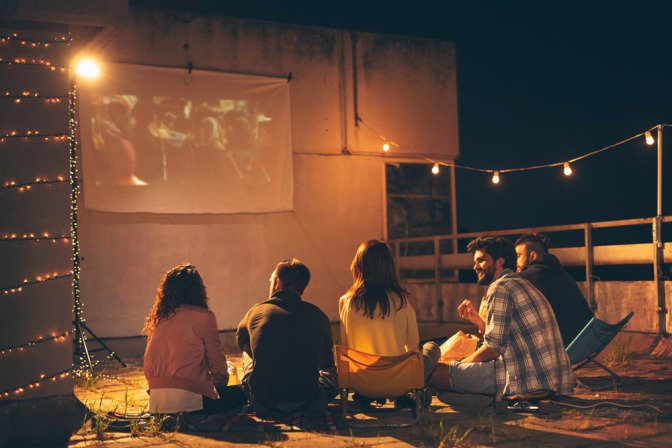 Eine Gruppe von Freunden sitzt an Silvester auf einem Dach vor einem selbstgebauten Outdoorkino.