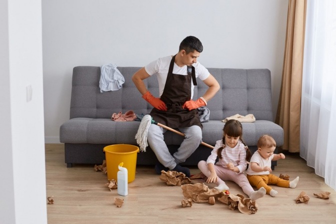 Mann reinigt seine Couch, während Kinder auf Boden sitzen und Ordnung stiften.