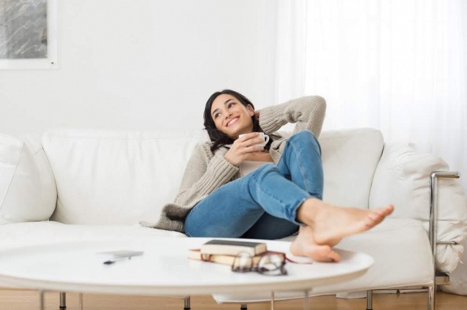 Eine Frau liegt auf einem Sofa und lächelt zufrieden. Sie hält eine Tasse in der Hand.