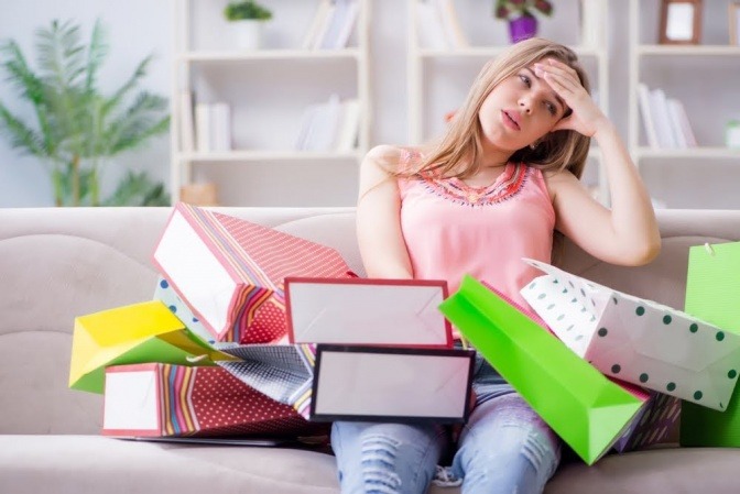 Eine Frau sitzt auf einem Sofa, auf ihrem Schoß und um sie herum liegen diverse Einkaufstüten. Sie wirkt resigniert und antriebslos.