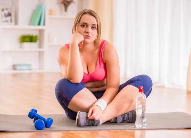 Eine Frau sitzt im Schneidersitz auf einer Iso-Matte in einem Fitnessraum. Mit einem Arm stützt sie Ihren Kopf und blickt demotiviert und lustlos zur Seite. 