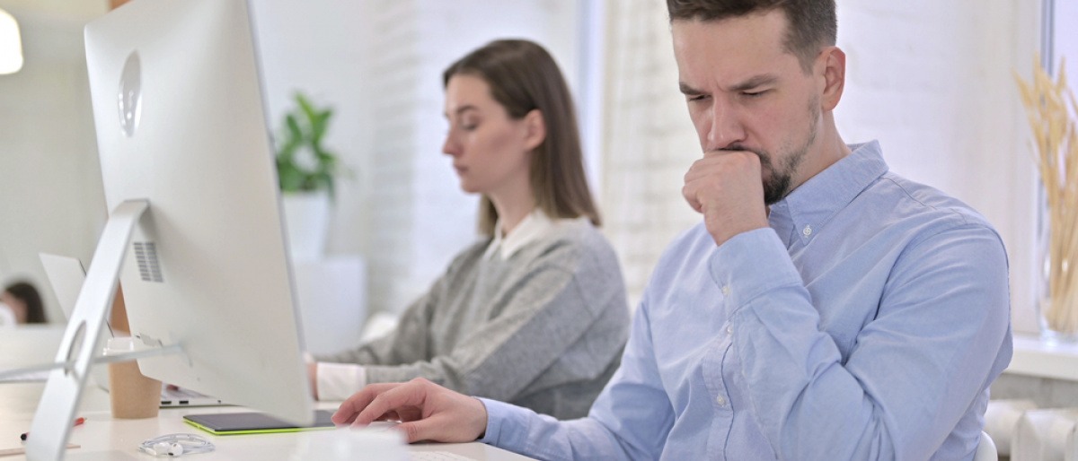 Neben einer im Büro sitzenden Frau hustet ein Mann 