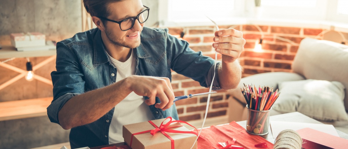 Geschenke originell verpacken Tipps - junger Mann verpackt Präsente auf Tisch.